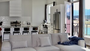 Une cuisine moderne avec des meubles blancs, un four encastré et une machine à café. On y trouve un coin repas avec une table blanche et des chaises noires. Un canapé gris est placé au premier plan, offrant une vue sur les montagnes par la fenêtre.