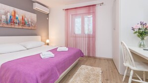Un dormitorio con cama, escritorio, silla, ventana con cortinas y un cuadro en la pared.