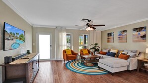 Un salon avec un plancher en bois, un canapé, deux fauteuils, une table basse, un téléviseur et un ventilateur de plafond.