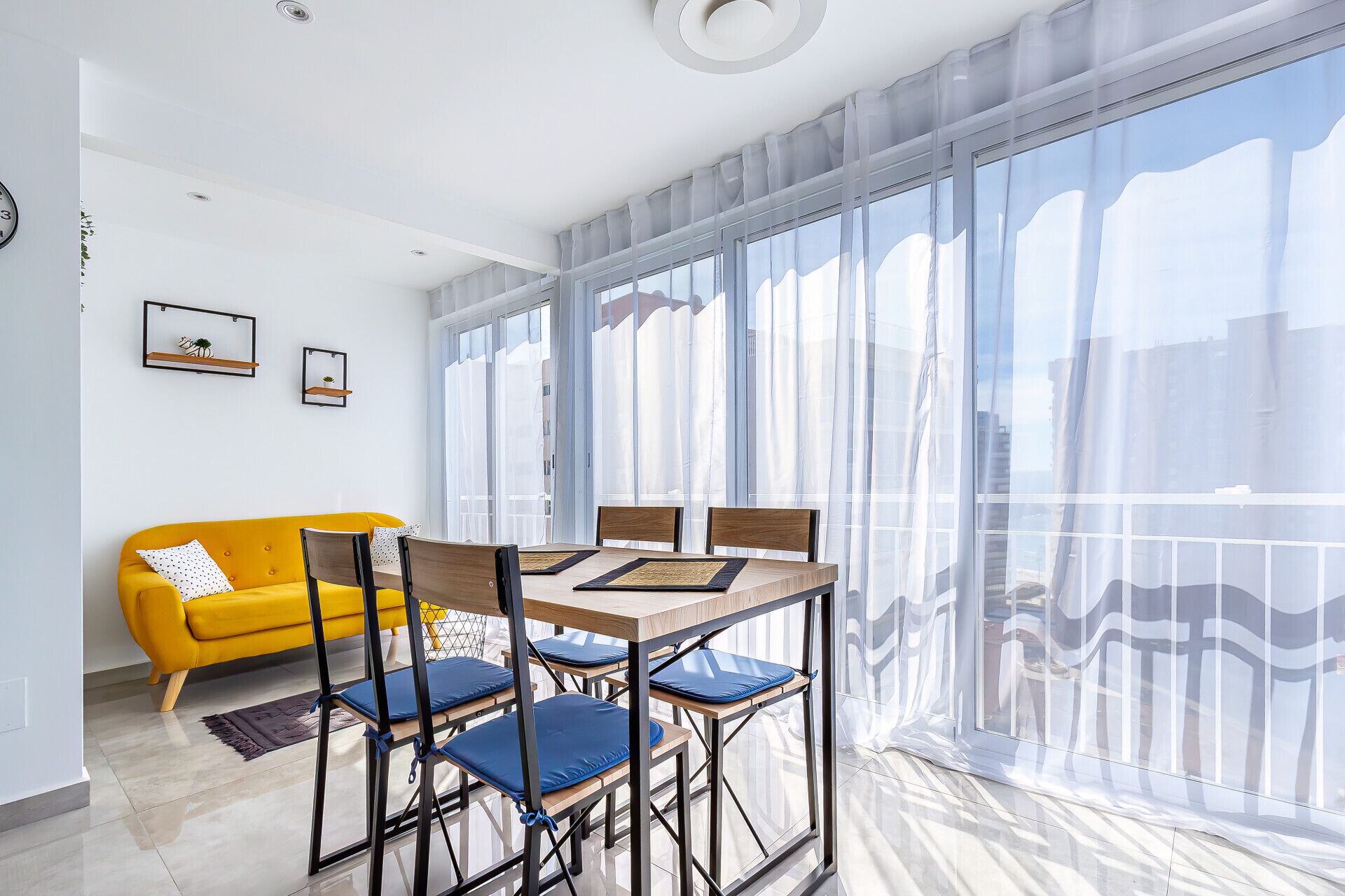 A modern living room with a yellow sofa, a dining table with chairs, and large windows with sheer curtains.