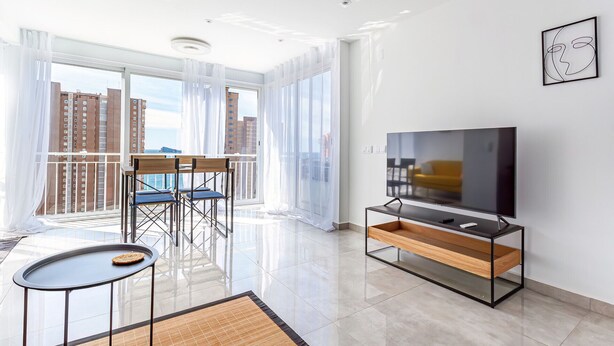 Ein modernes Wohnzimmer mit großem Fernseher, einem Essbereich mit Stühlen und einem Balkon mit Blick auf hohe Gebäude.