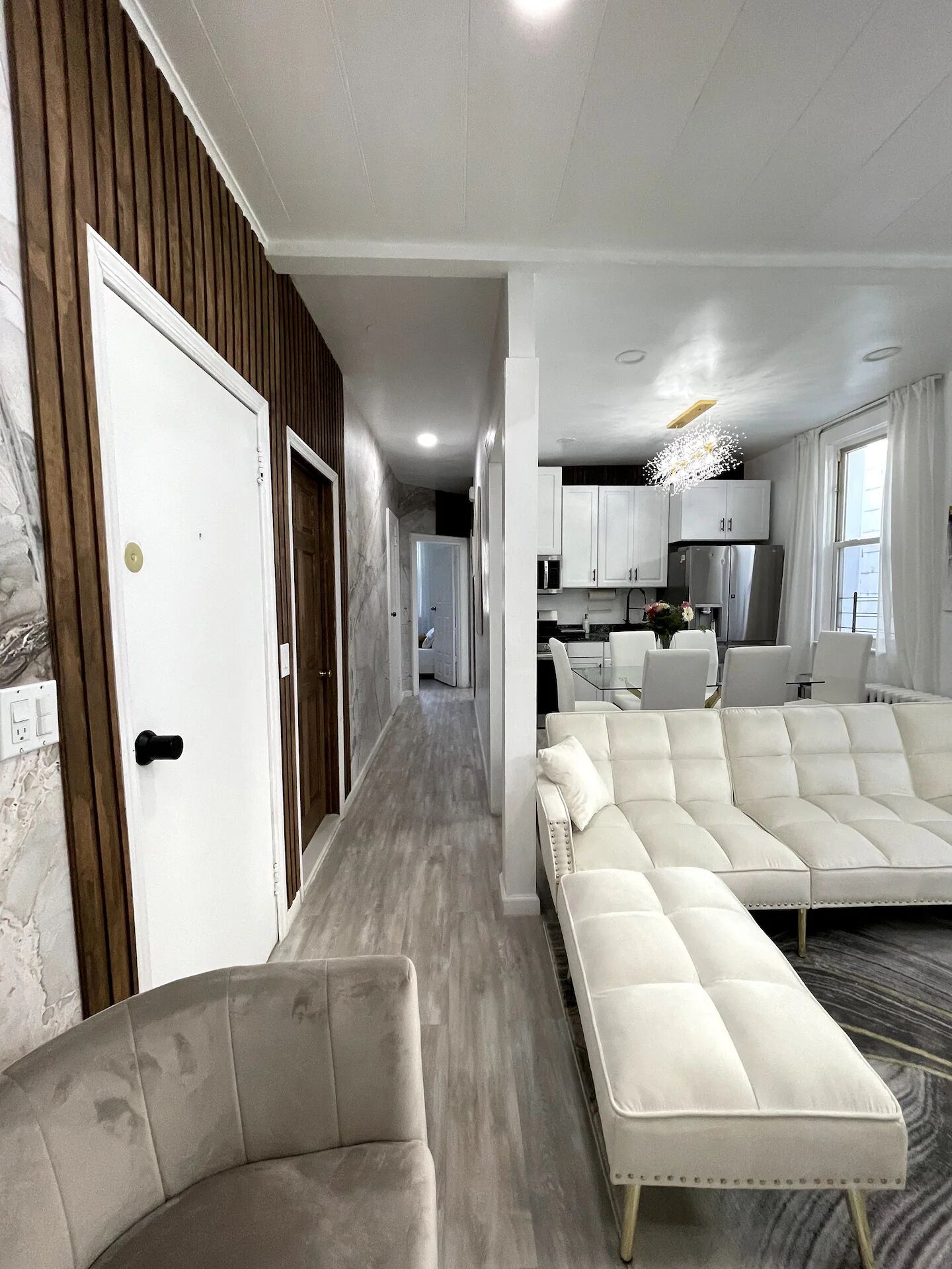 Una sala de estar moderna con un sofá y un sillón, una zona de cocina con armarios y electrodomésticos, y un pasillo que conduce a otra habitación.