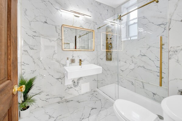 Un baño moderno con paredes de mármol, un lavamanos blanco, un espejo y una ducha con cerramiento de vidrio.