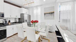 A modern kitchen with white cabinets, a stainless steel refrigerator, and a glass dining table with white chairs. - Luxury Living in the Heart of NY (Yonkers)