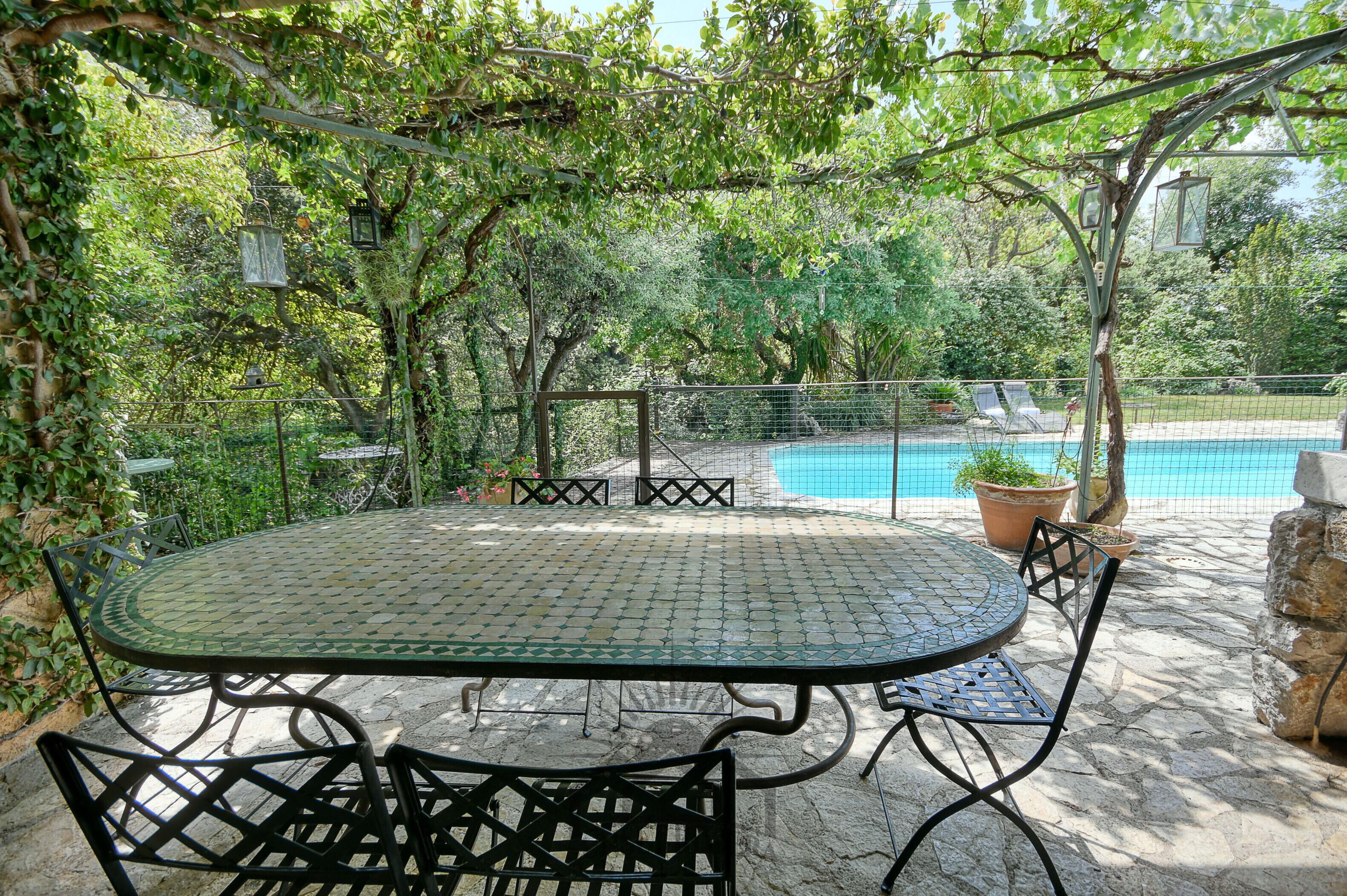 A patio with a round table and chairs, surrounded by a stone floor and a pool in the background.