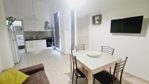 Una cocina moderna con armarios blancos y horno empotrado, un comedor con una mesa de vidrio y cuatro sillas, y un televisor de pantalla plana montado en la pared.