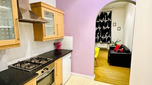 Een compacte keuken met een gasfornuis, oven en een kleine koelkast. De keuken heeft houten keukenkastjes en een zwart aanrecht. Een paarse wand en een deurpost leiden naar een zithoek met een zwarte bank en een rode kussen.