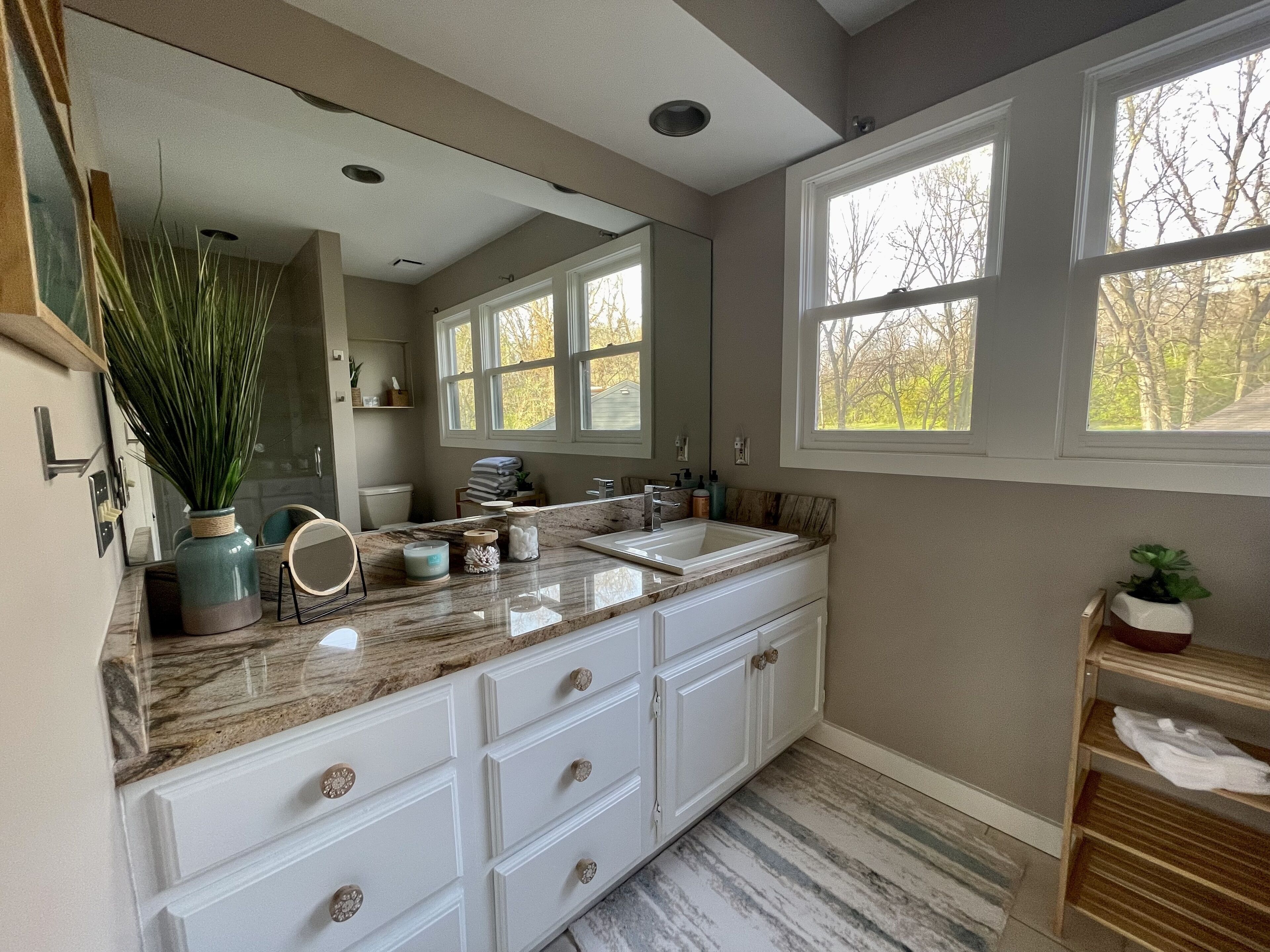 A modern bathroom with a large vanity, a sink, and a mirror. There are windows providing natural light and a view of trees outside.