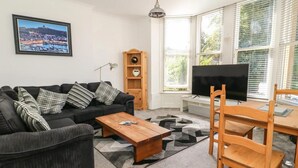Sala de estar con un sofá negro, una mesa de centro de madera y un televisor. También hay una estantería y una ventana con persianas.