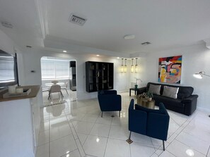 Un salon moderne avec un canapé noir, des fauteuils bleus et une table basse. On y trouve également un coin repas avec une table et des chaises blanches, ainsi qu’un coin cuisine avec un meuble noir et une fenêtre équipée de stores.