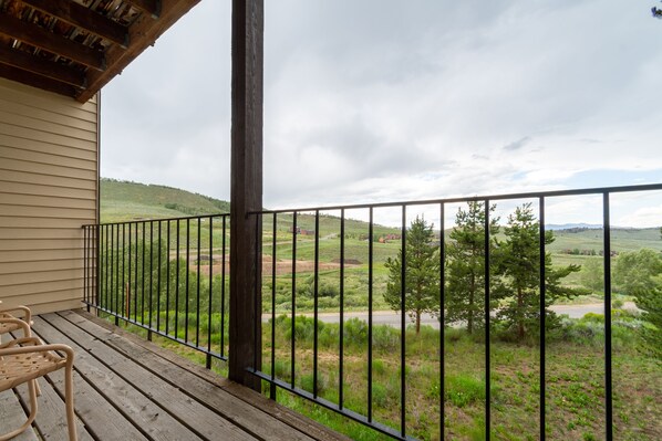 A wooden deck with metal railings overlooking a grassy landscape with trees and a road. - Granby Condo with Private Hot Tub (Granby)