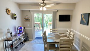 Uma sala de jantar com mesa posta para seis lugares, um ventilador de teto, uma TV fixada na parede e vista para o exterior por uma porta de vidro.