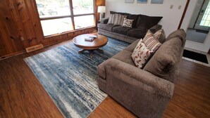 A living room with a brown sofa, a round coffee table, and a blue patterned rug. There are three framed pictures on the wall and a window with a view of the outdoors. - Streamside Family Friendly Accommodations minutes from Ohiopyle! (Farmington)