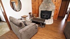 A cozy living room with a stone fireplace, wooden walls, a grey sofa, and a small wooden table. - Streamside Family Friendly Accommodations minutes from Ohiopyle! (Farmington)