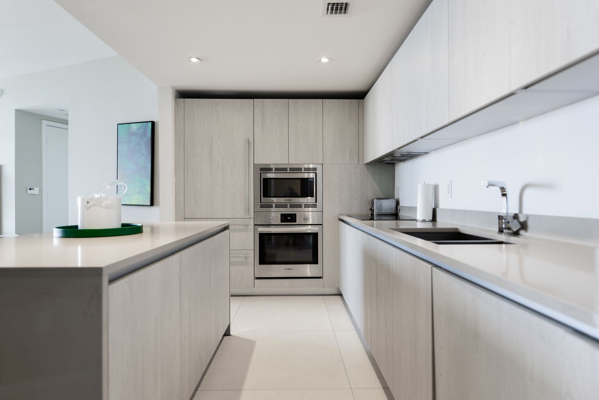 A modern kitchen with a central island, built-in ovens, a microwave, and a stainless steel sink.