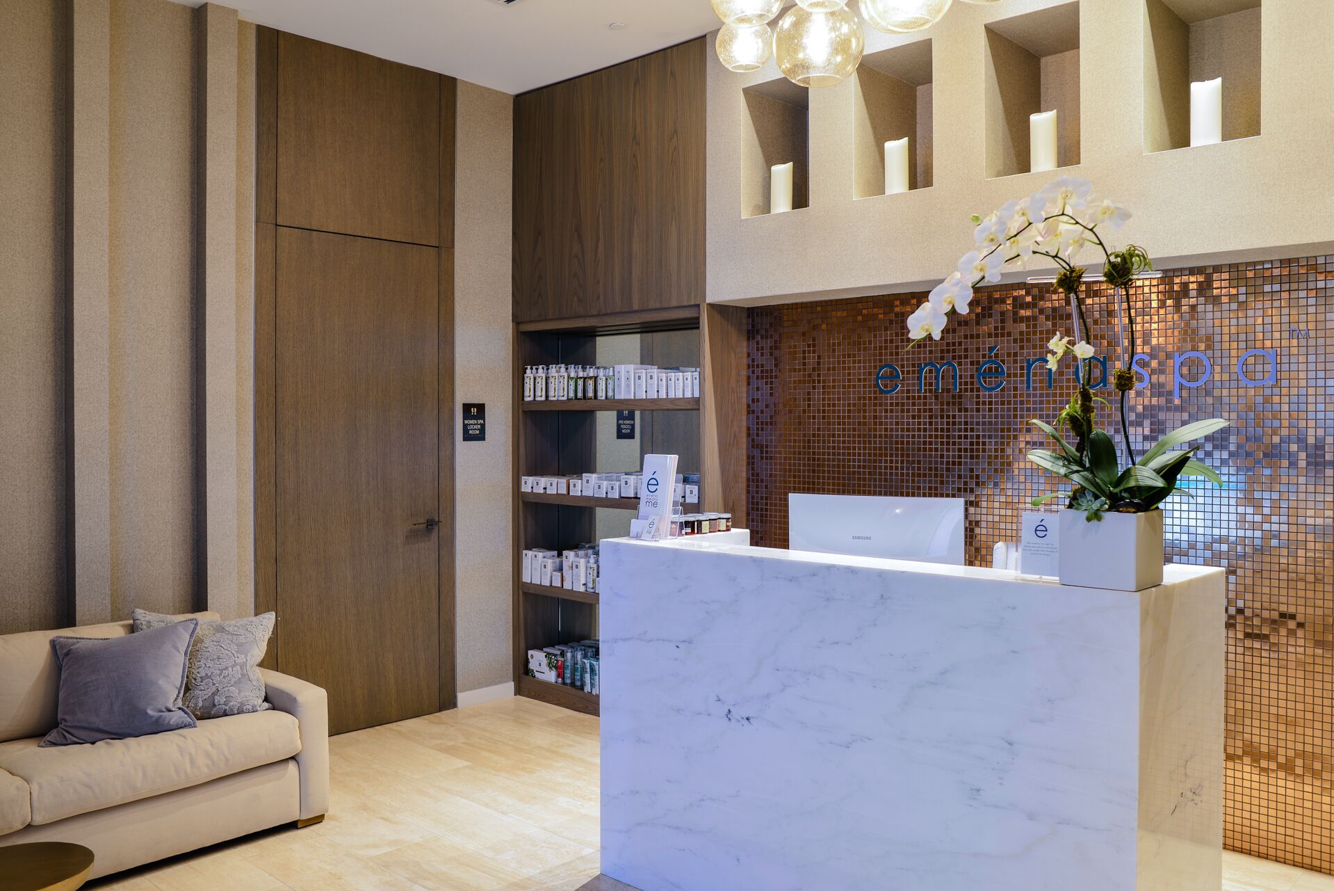 A reception desk with a white marble top, a sofa with cushions, a shelf with products, and a wall with a mosaic pattern and orchids.