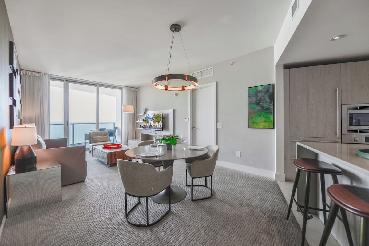 A modern hotel room with a dining area, a kitchen, and a balcony with a view.