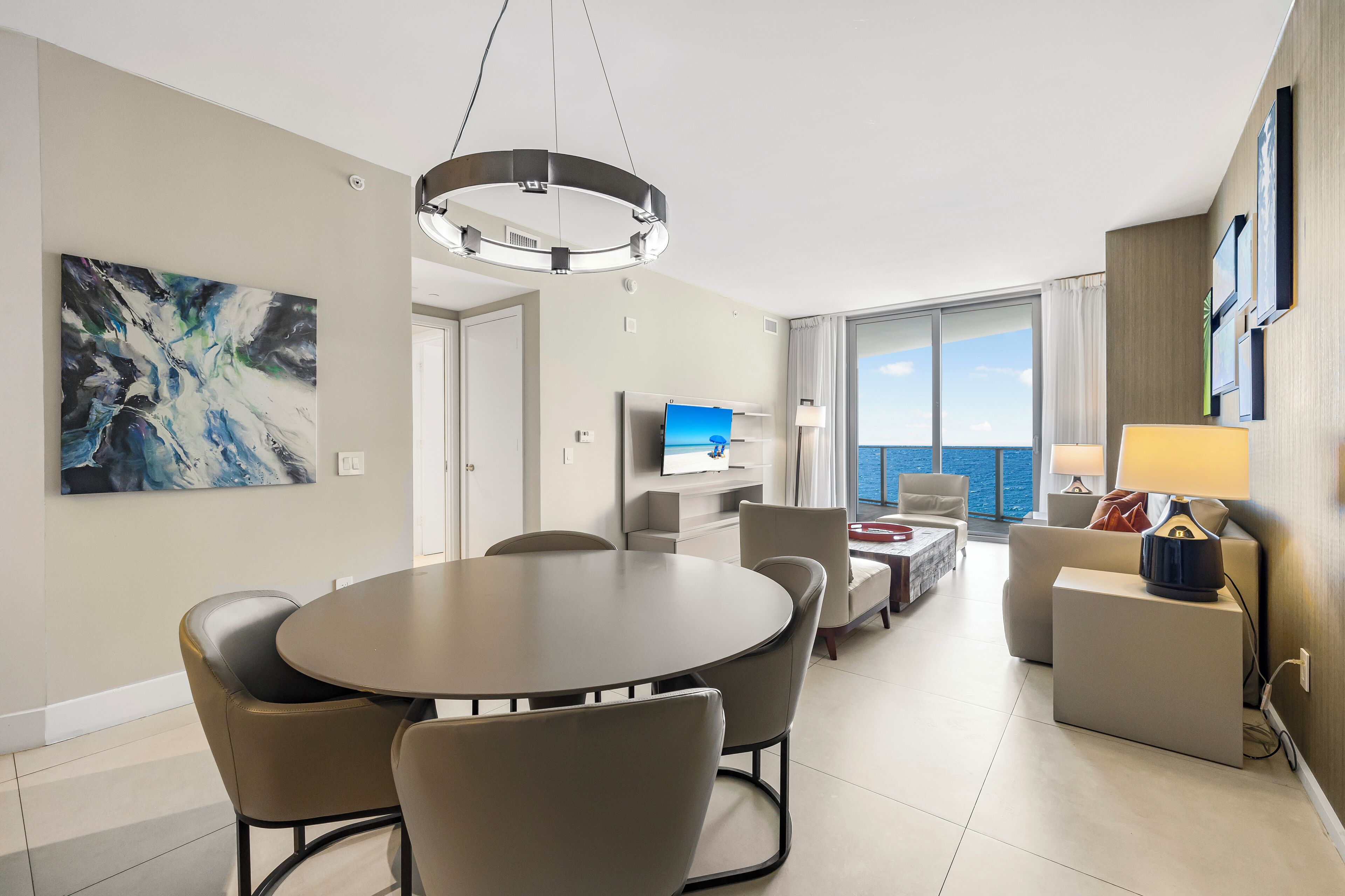 A modern hotel room with a dining area, a flat-screen TV, a large window with a view, and a painting on the wall.