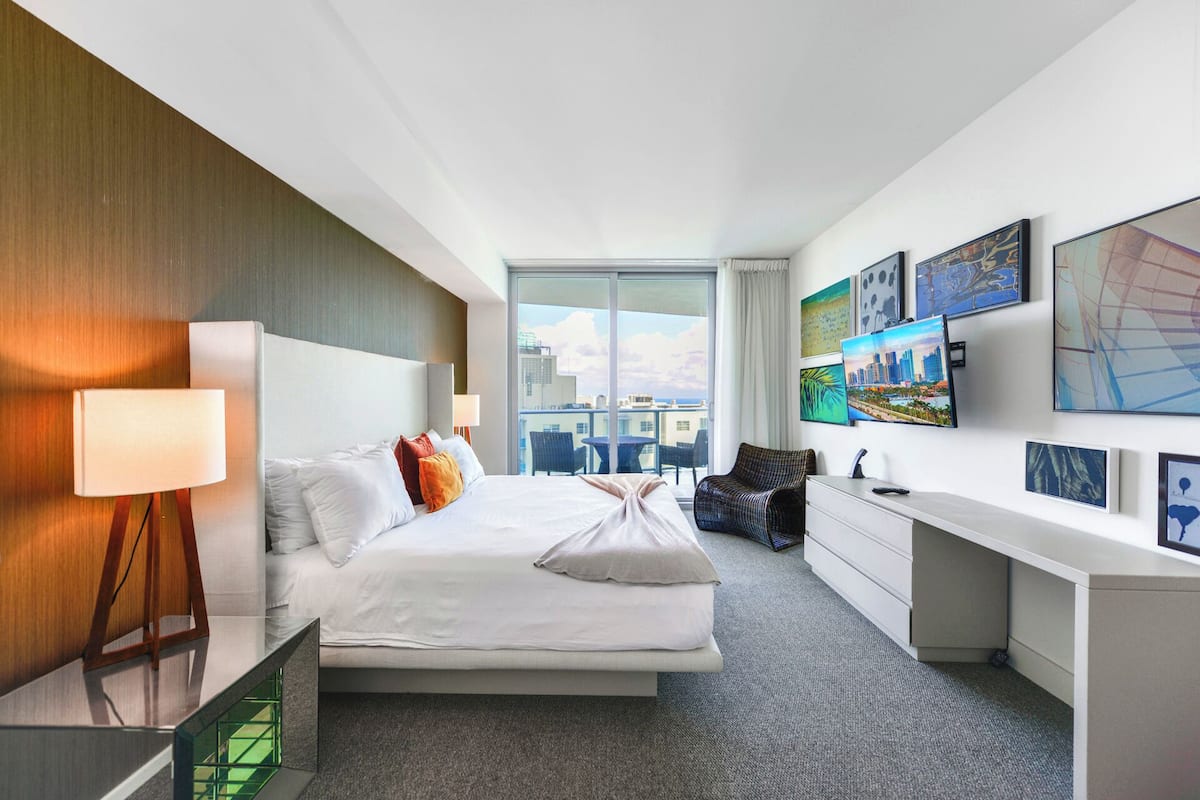 A modern hotel room with a large bed, a desk, and a view of the city through a large window.