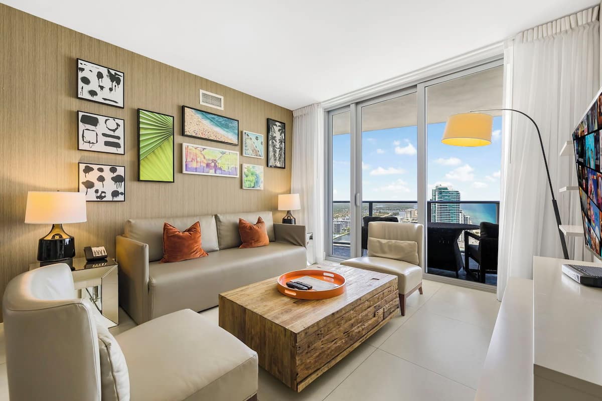 A modern hotel room with a large balcony, a sofa set, a coffee table, and various wall art.