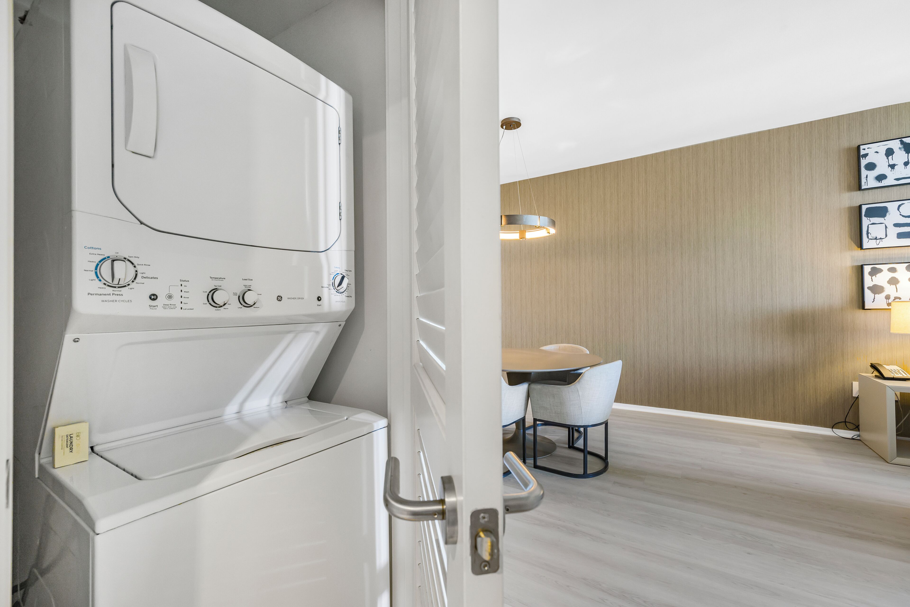 A modern hotel room with a built-in washer and dryer, a wooden wall, a dining area with a table and chairs, and a small desk with a telephone.