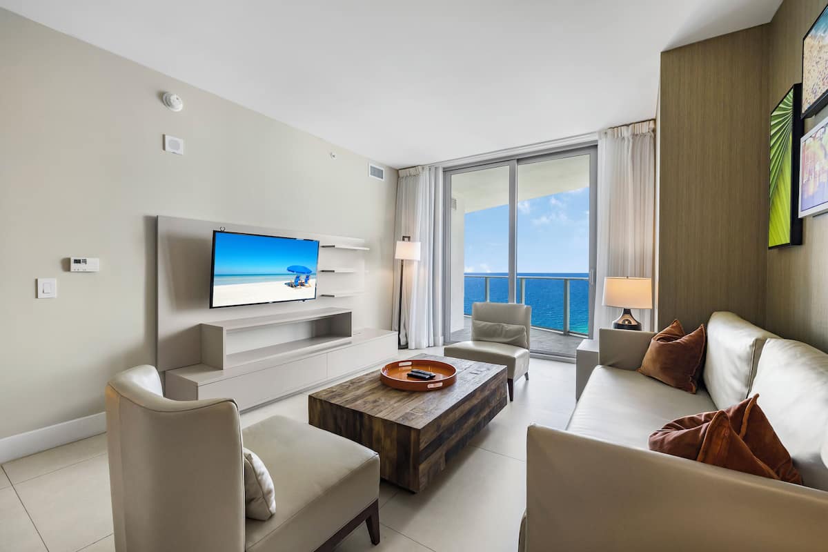 A modern hotel room with a flat-screen TV, comfortable seating, a wooden coffee table, and a large window offering a view of the ocean.