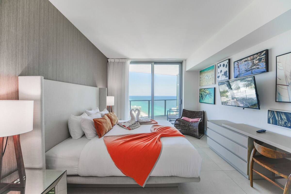 A modern hotel room with a large bed, a desk, and a view of the sea.