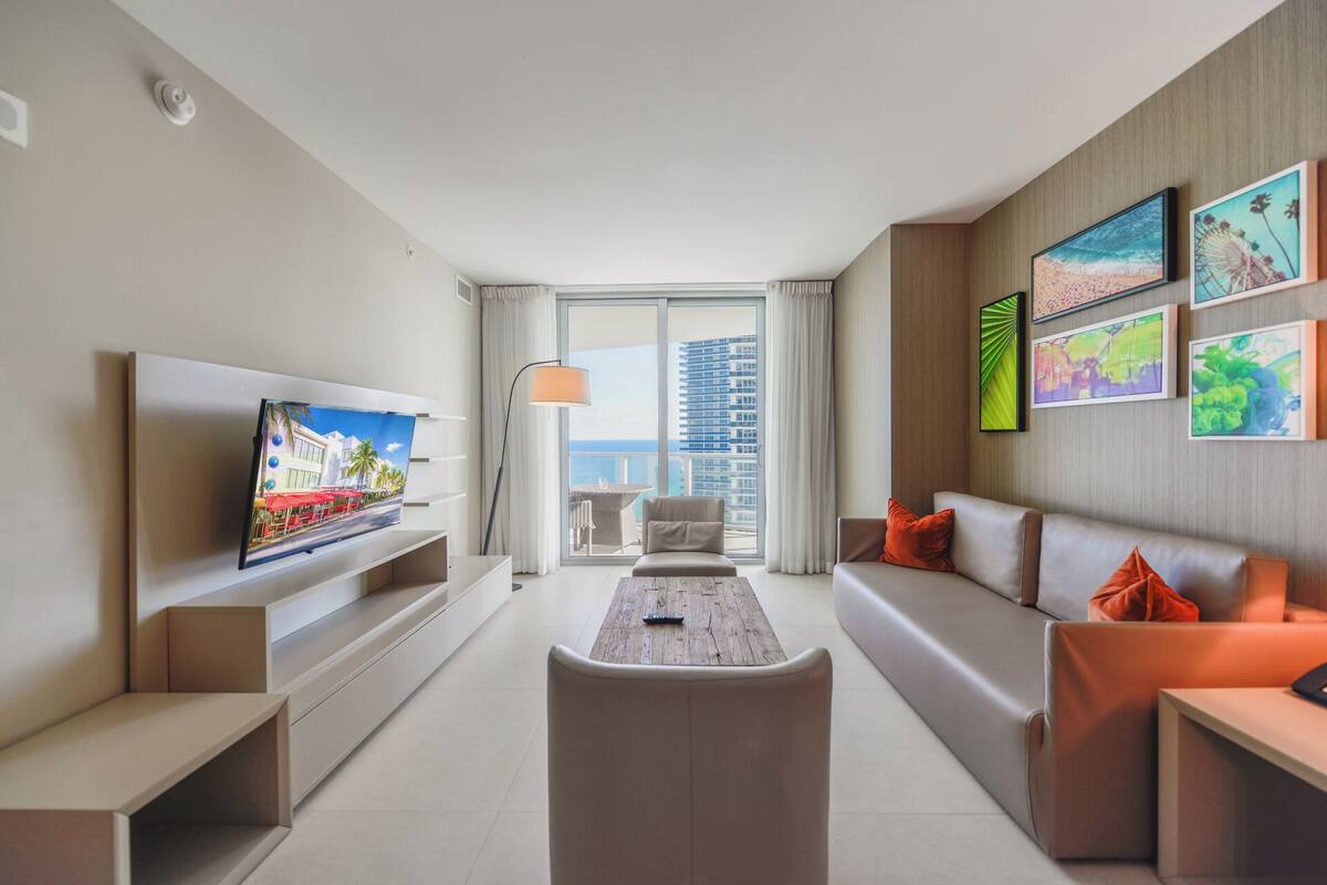 A modern living room with a large TV, a sofa, a coffee table, and a view of the cityscape through large windows.
