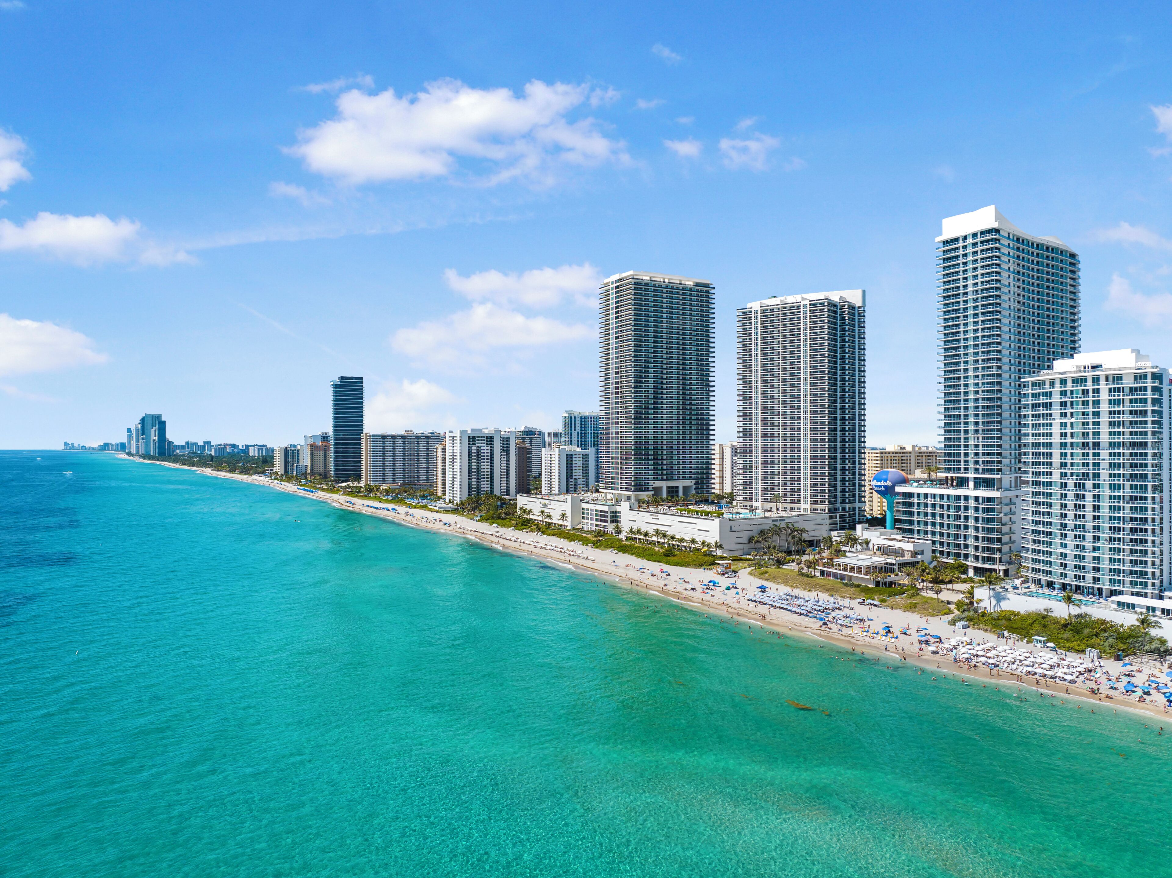 A coastal cityscape with high-rise buildings, a beach, and clear turquoise waters.