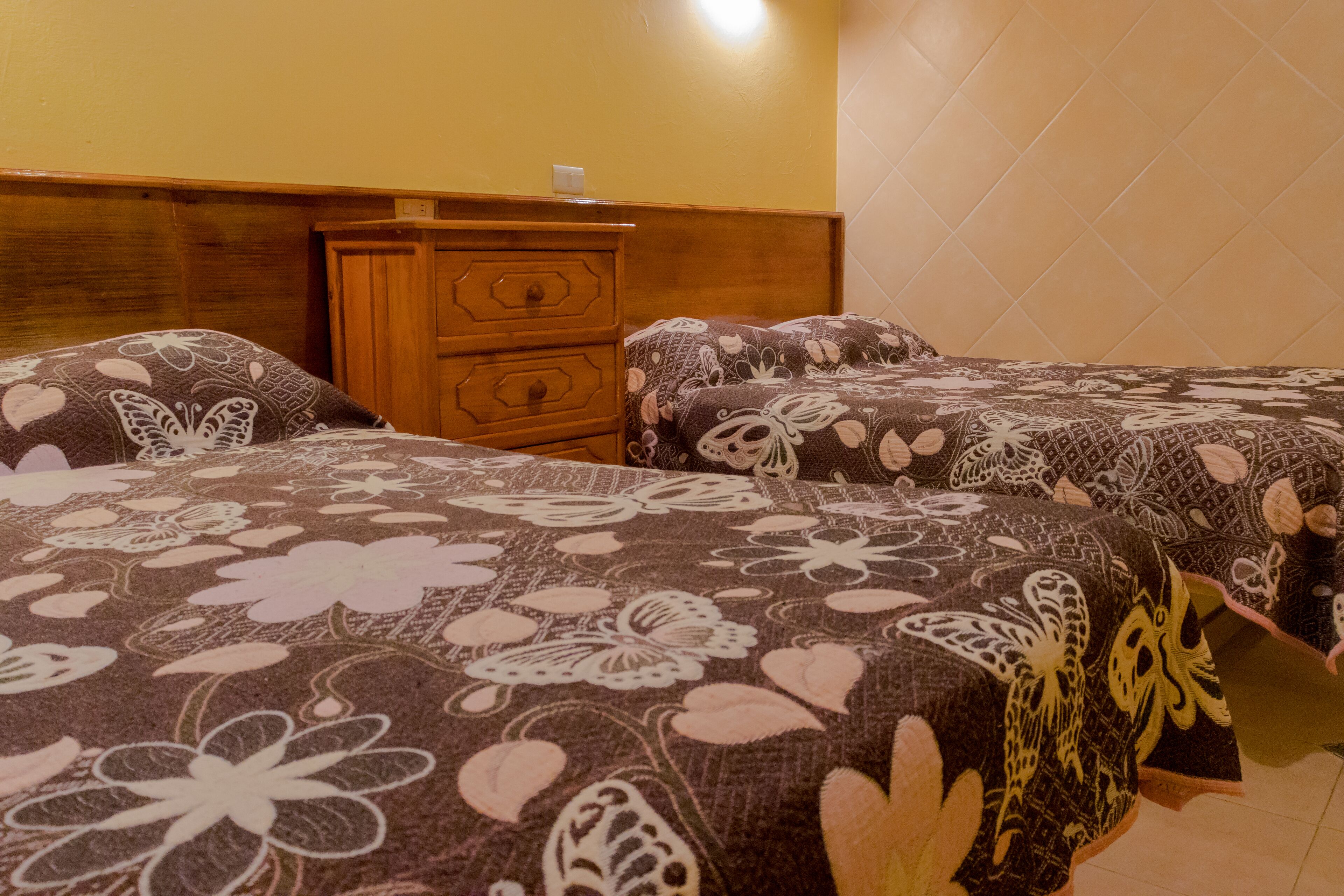 A hotel room with two beds, each with floral-patterned bedding, a wooden dresser, and a wall-mounted light.