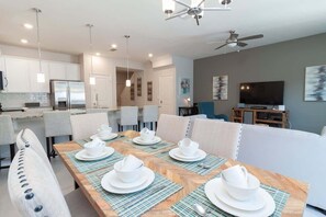 Un comedor moderno con una larga mesa de madera dispuesta para seis comensales, sillas blancas y una cocina al fondo.