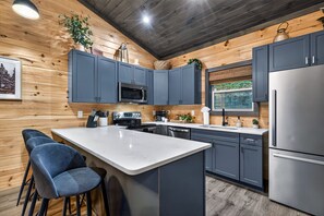 A modern kitchen with grey cabinets, a stainless steel refrigerator, a microwave, and a large island with bar stools. - Modern Couples Cabin Mountain Retreat near Gatlinburg (Gatlinburg)