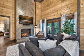 A cozy living room with a fireplace, wooden walls, a sofa, and a large window. - Modern Couples Cabin Mountain Retreat near Gatlinburg (Gatlinburg)