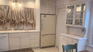 Une cuisine avec des meubles blancs, un réfrigérateur, une fenêtre avec des rideaux et une table à manger avec des chaises.