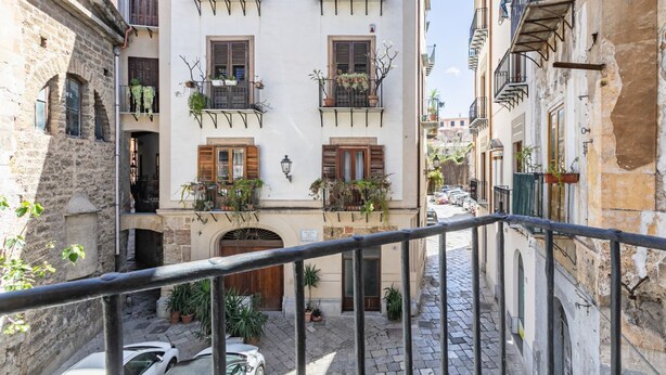 Une rue étroite, bordée de bâtiments des deux côtés, avec des balcons ornés de plantes et une voiture garée le long de la rue.