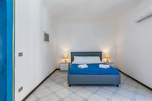 A bedroom with a bed, two bedside tables with lamps, a wall-mounted air conditioner, and a small framed picture on the wall.
