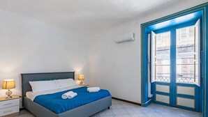 Ein Schlafzimmer mit einem Bett, zwei Nachttischen, einem Fenster mit blauen Rollläden und einer Tür mit Blick auf Gebäude.