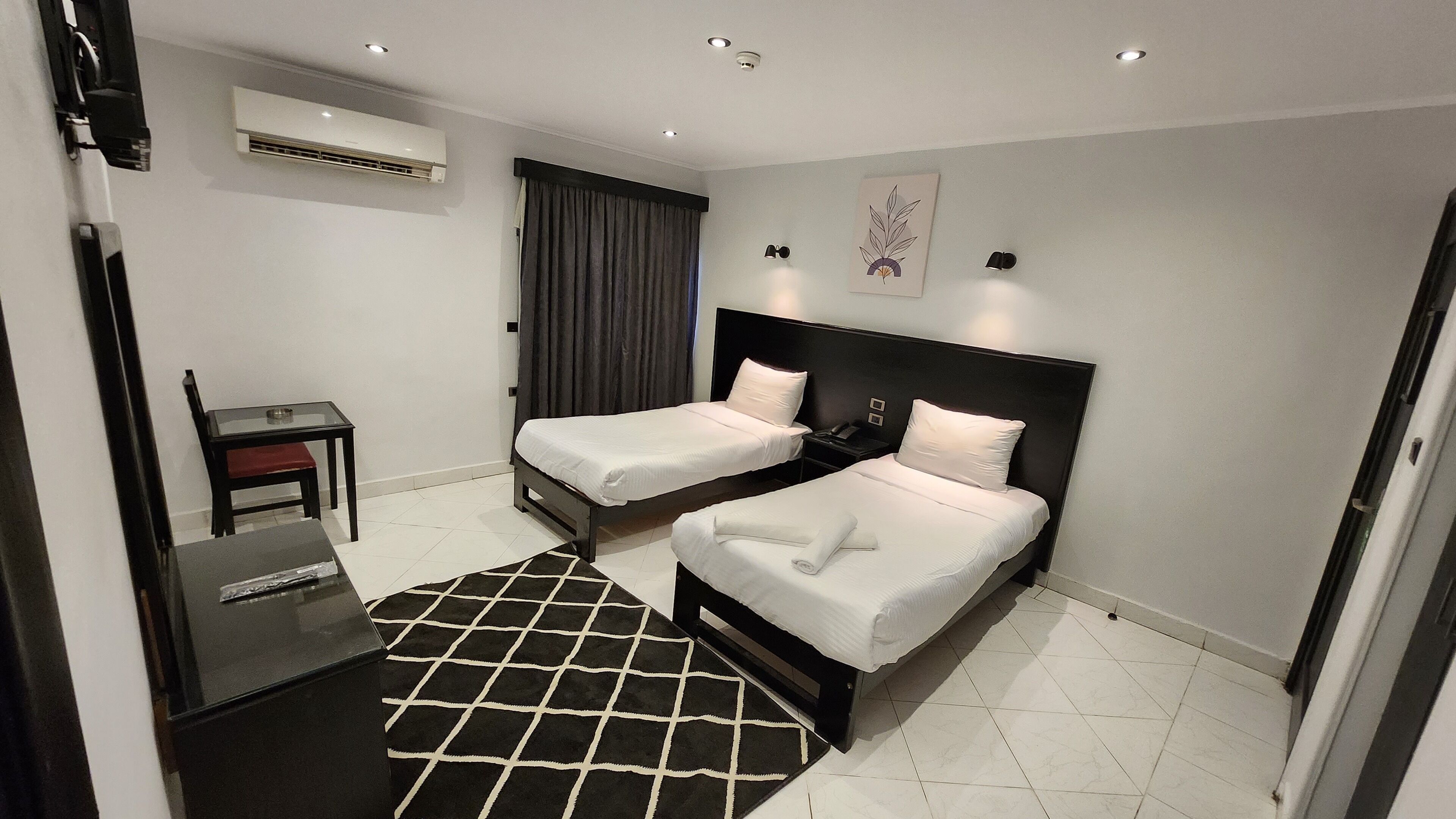 A hotel room with two single beds, a desk, a chair, a black and white rug, and an air conditioning unit on the wall.