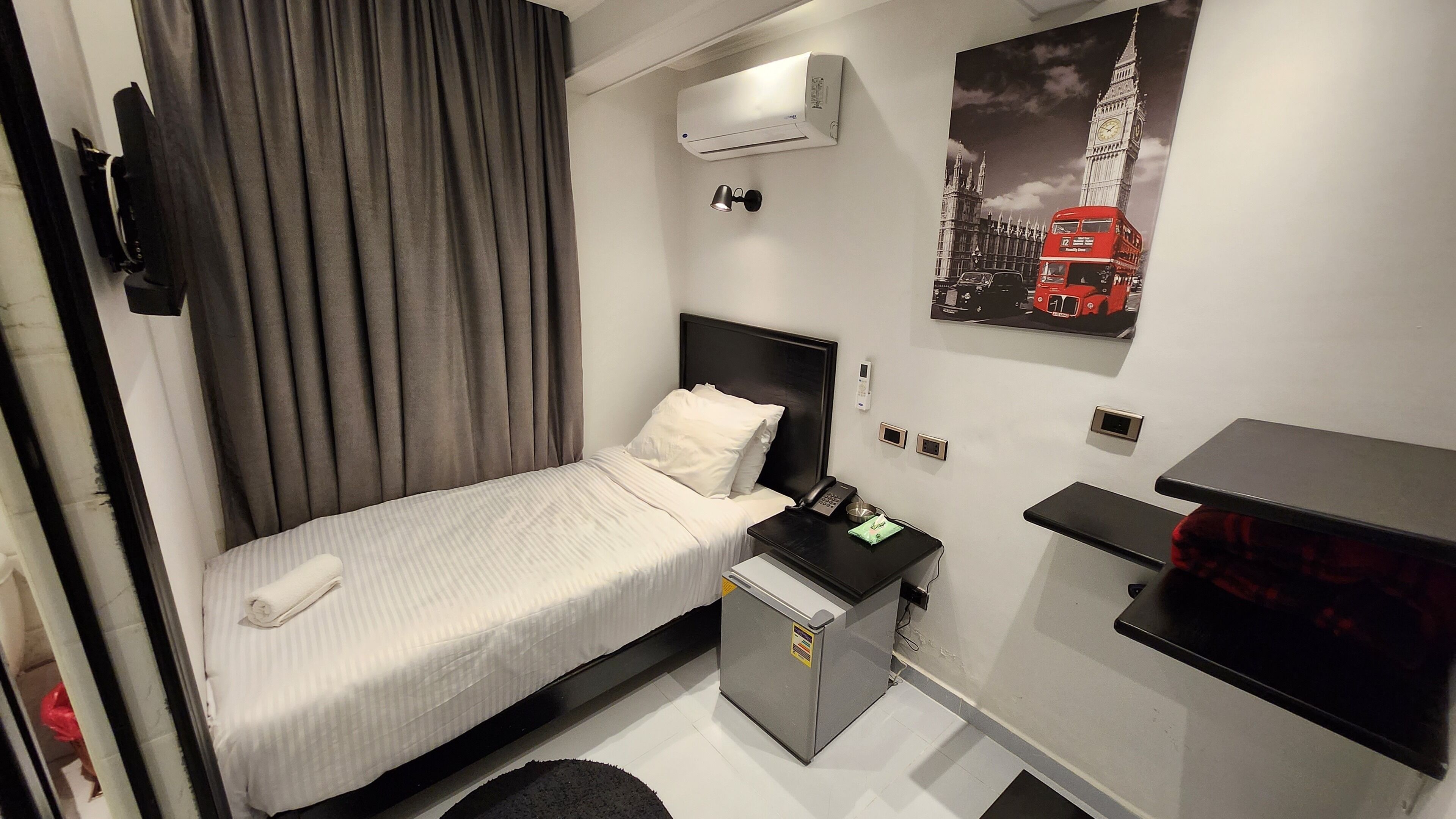 Habitación de hotel compacta con cama individual, escritorio pequeño, mini refrigerador, aire acondicionado instalado en la pared, teléfono y un cartel de un autobús rojo de dos pisos.