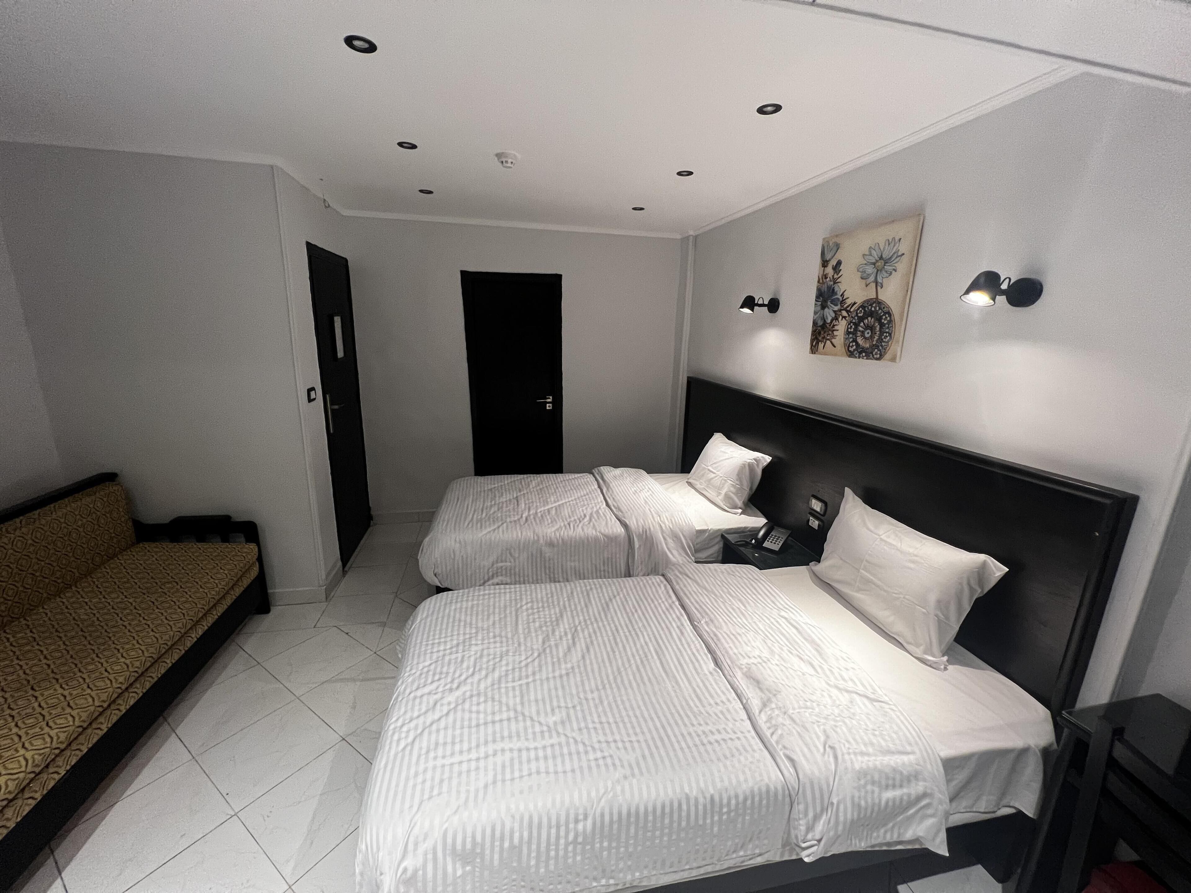 Habitación de hotel con dos camas, un sofá, una mesita de noche con lámpara y una obra de arte en la pared.