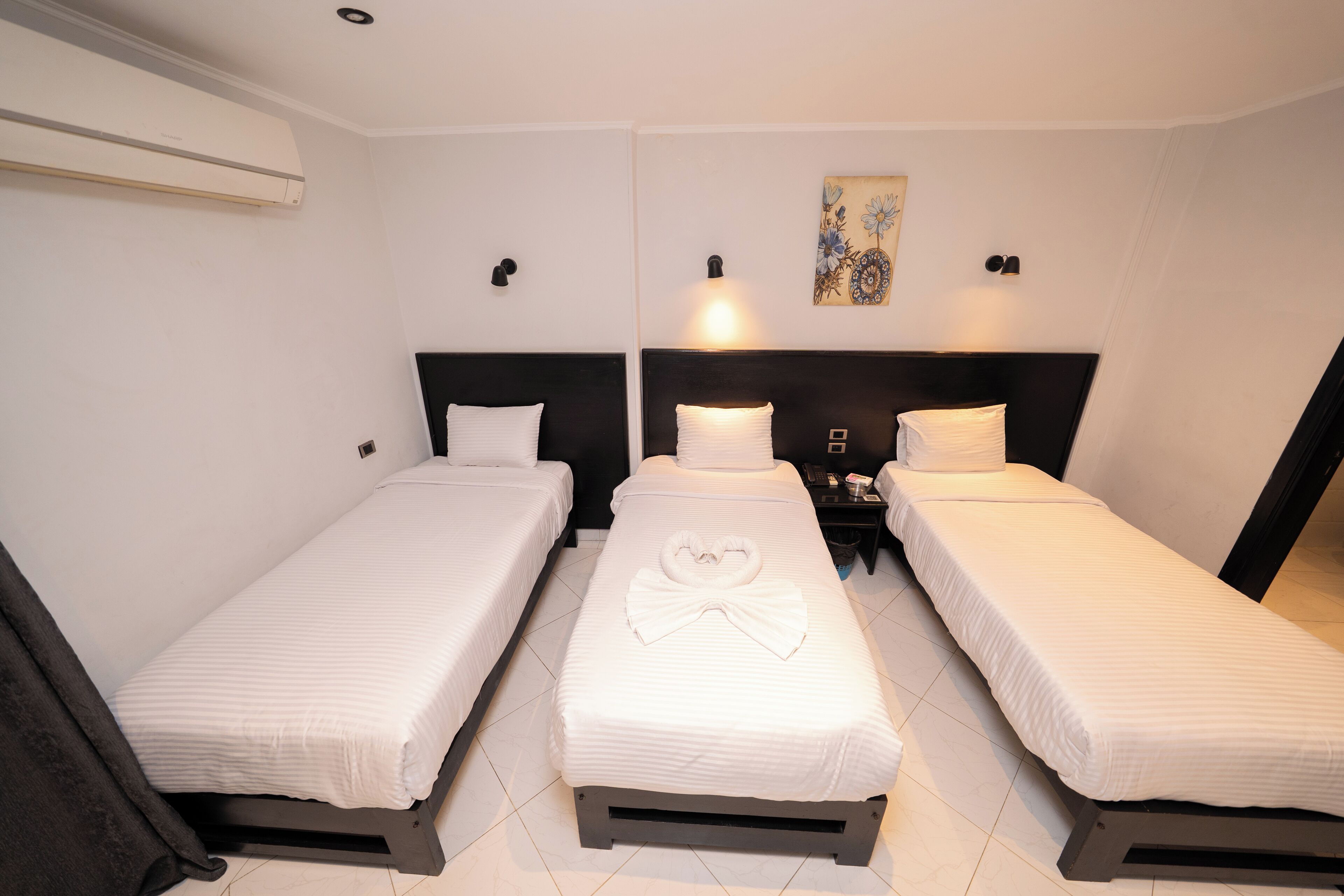 Une chambre d’hôtel avec trois lits simples, chacun agrémenté de draps blancs et d’une serviette en forme de cœur posée sur le lit du milieu. On y trouve deux tables de chevet fixées au mur, avec des lampes, et un tableau au-dessus du lit du milieu.