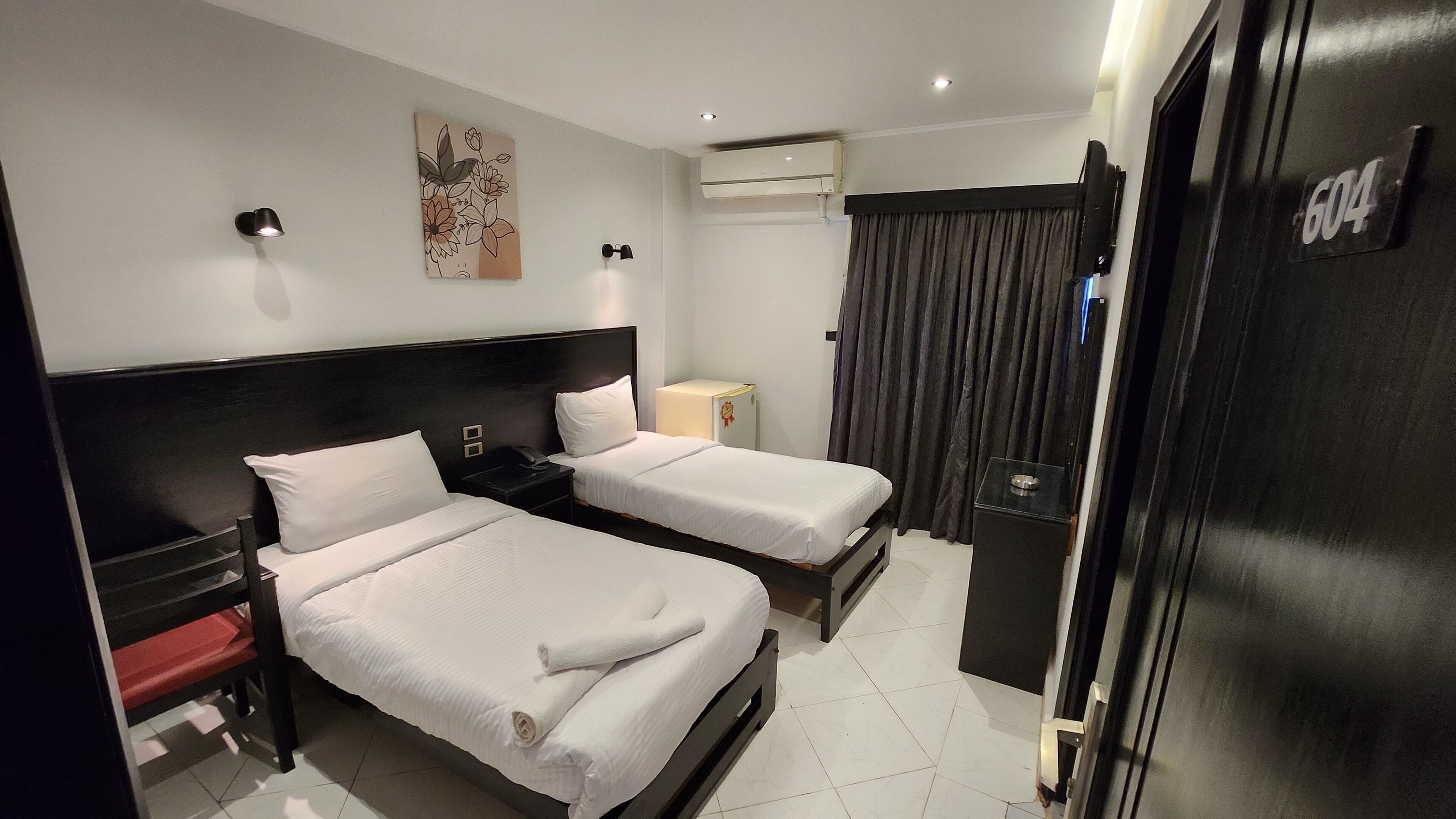 Habitación de hotel con dos camas, una mesita de noche, una pequeña mesa, un equipo de aire acondicionado y una puerta con un número.