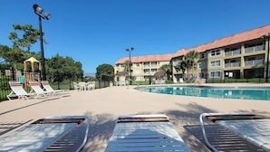 Un espace aménagé au bord de la piscine, avec des chaises longues, un kiosque et un bâtiment doté de balcons.