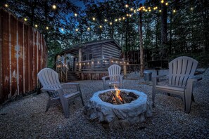 Un espace extérieur rustique aménagé avec des chaises Adirondack disposées autour d’un foyer, une terrasse en bois et des guirlandes lumineuses, le tout dans un cadre boisé.