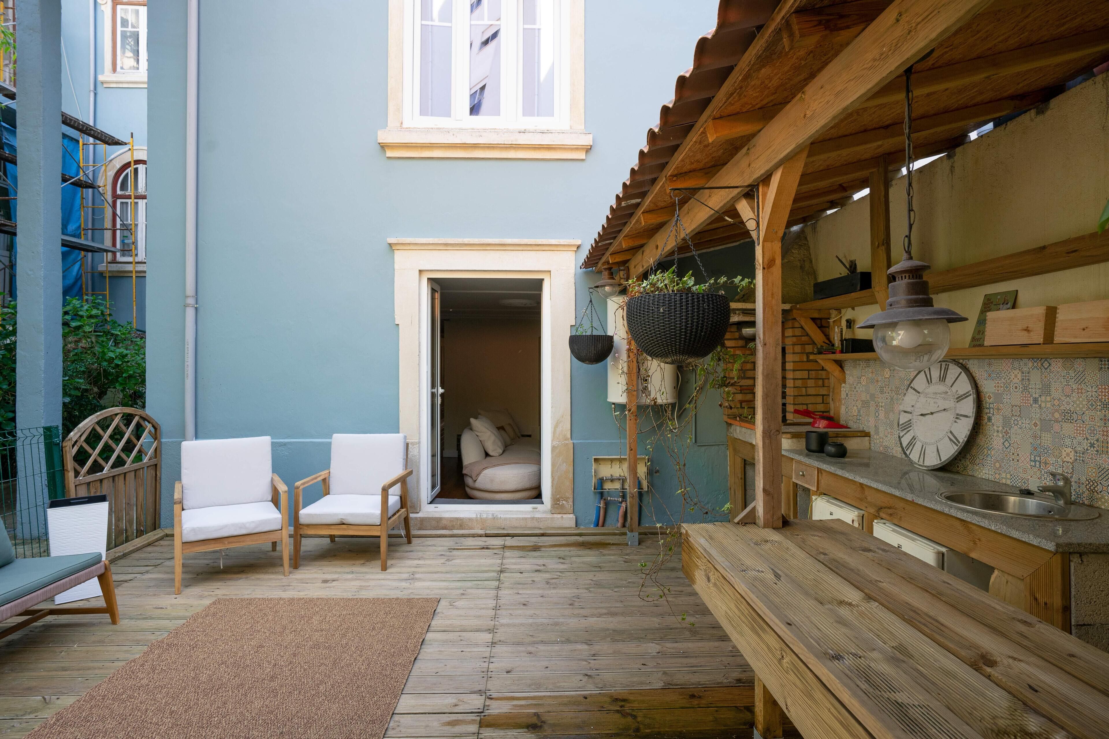 Een houten terras met buitenmeubilair, een klein keukentje met een klok, en een deur die leidt naar een slaapkamer.