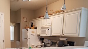 Eine Küche mit weißen Schränken, einer Mikrowelle, einem Toaster und einem Kühlschrank. An der Kochinsel stehen zwei Holzstühle.