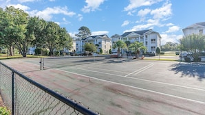 Ein Tennisplatz mit Maschendrahtzaun, umgeben von Bäumen und einem Gebäude mit mehreren Balkonen.