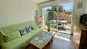 Un salon avec un canapé vert, une table basse en bois et une porte-fenêtre coulissante donnant sur une terrasse extérieure avec un mur en pierre et une table.