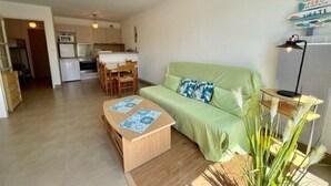 Un espace de vie compact avec un canapé vert, une table basse en bois et une petite table d’appoint avec une lampe. On aperçoit un coin cuisine au fond.