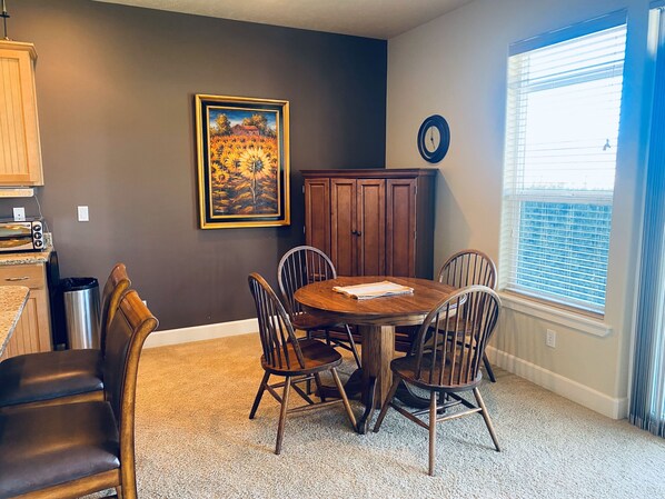 Un coin repas avec une table ronde, quatre chaises, un tableau au mur, une horloge et une fenêtre avec des stores.